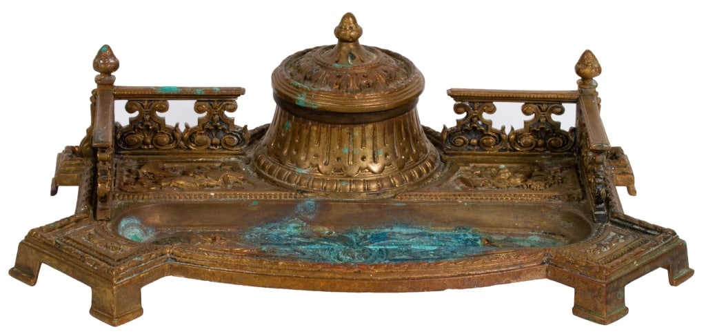Gilt Brass Inkwell with Pen Tray: Item/Description: Gilt brass inkwell pierced back gallery, arched front with a pen holder, covered center inkwell with glass insert. Floral and musical instrument designs. Stamped Germany. Material: G