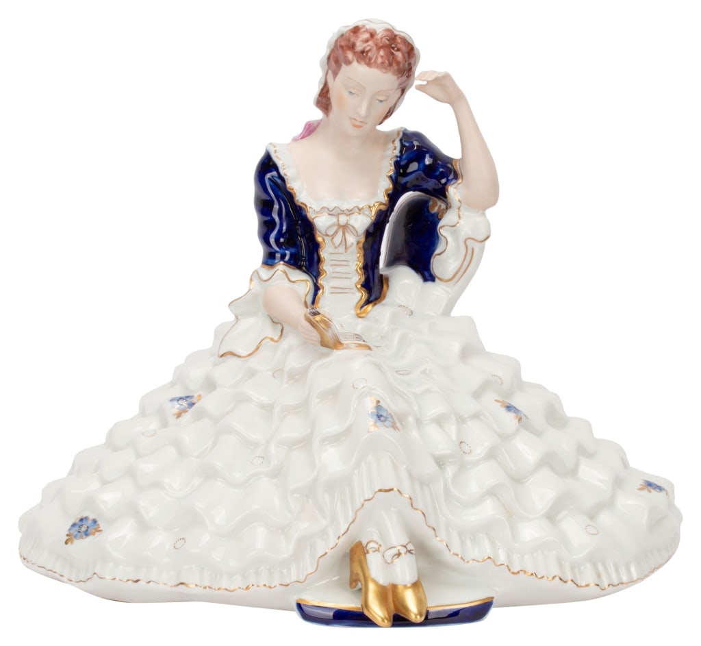 Royal Dux Seated Lady Reading Porcelain Figurine: Item/Description: Royal Dux figurine of a woman in a crinoline dress with cobalt blue accents. Seated, reading a book, she has both a bisque and glazed finish with hand painted with gilt details. Make