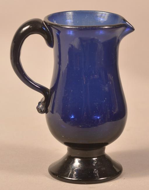 Stiegel Type Cobalt Flint Glass Footed Cream Pitcher.: Stiegel-Type Cobalt Flint Glass Footed Cream Pitcher. Blown with comet tail and applied loop handle. 4-1/4" high. Condition: Very good.