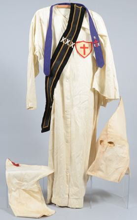 Vintage Ku Klux Klan Uniform, front buttoned whit: Vintage Ku Klux Klan Uniform, front buttoned white robe, attached short white cape with a sewn-on shield and cross patch, two hoods (one closed face and one open face), and two various sashes, circa.