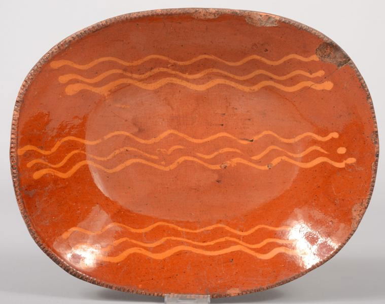 Redware Dished Platter with Slip Decoration. Oblong: Redware Dished Platter with Slip Decoration. Oblong dish has three sets of three yellow wavy lines. Coggle rim. 15 1/2"l. X 12 1/2"w. X 2 3/4"d. Condition: Two small chips and four larger chips off ri