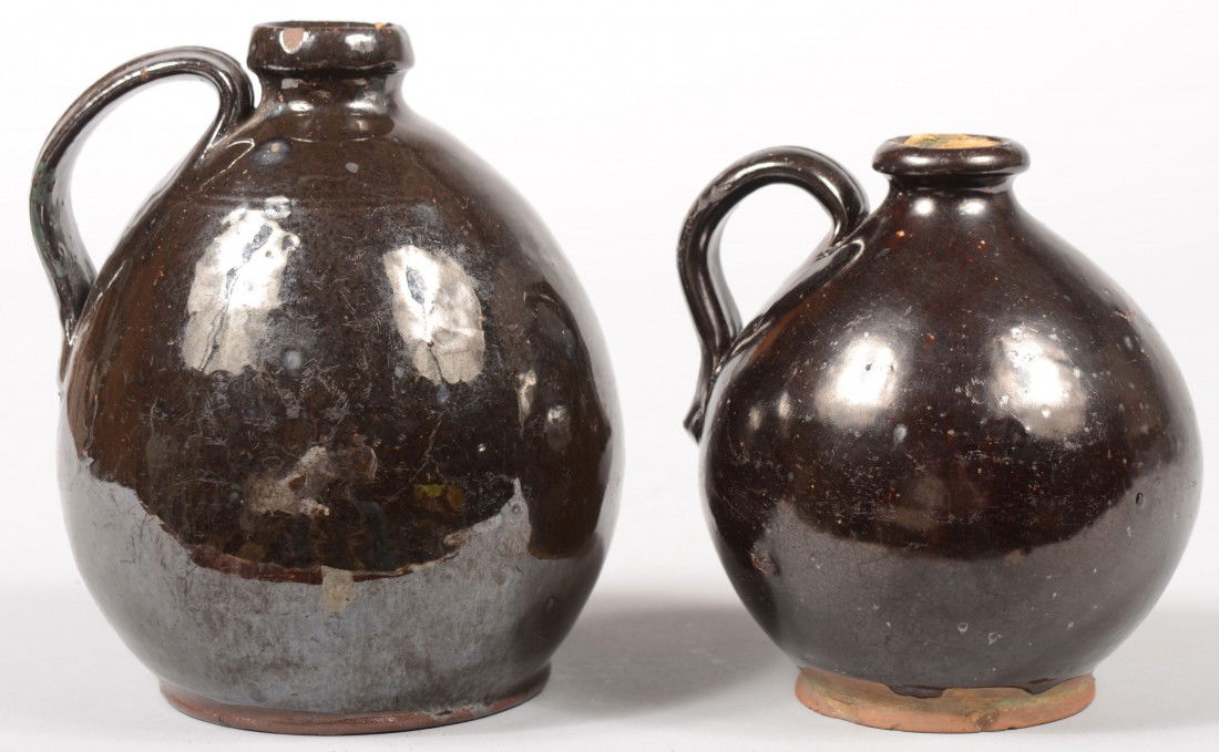 Two Similar Manganese Glazed Redware Jugs. Bulbous: Two Similar Manganese Glazed Redware Jugs. Bulbous ovoid bodies with bead foot, ring spout and applied strap handle. 5 1/4"h. & 6 1/4"h. Condition: Both good. Larger has small chip on spout rim.