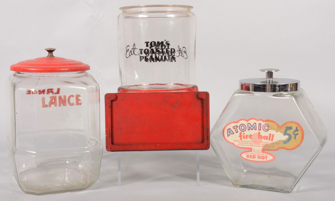 Three Branded Glass Product Containers. A cylindri: Three Branded Glass Product Containers. A cylindrical jar bearing: "Eat Tom's Toasted Peanuts" sitting on a red stamped steel sign holder. 8 3/4"h. X 9 1/2"d.; a diamond shaped jar with chromed lid an