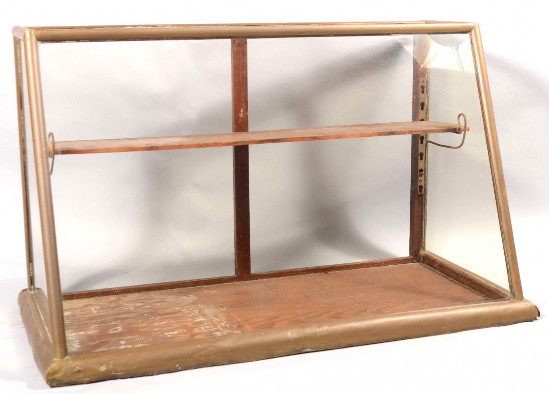 Wedge Shaped Glass Counter Top Display Case. Label: Wedge Shaped Glass Counter Top Display Case. Labeled: "Charles C. Geissler, Maker, Phila., PA." Brass sheet over wood frame with wood base. Glass on front, sides, and top. 24"h. X 40"w. X 18 1/2"d. ba