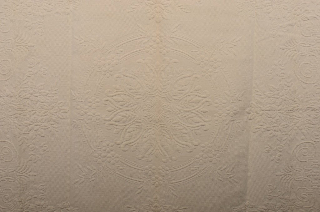 White Cotton Machine Trapunto Bedspread. Circular: White Cotton Machine Trapunto Bedspread. Circular central medallion surrounded by a scrolling floral border all on a field of small outlined squares. The hemmed head and foot edges have woven in ident