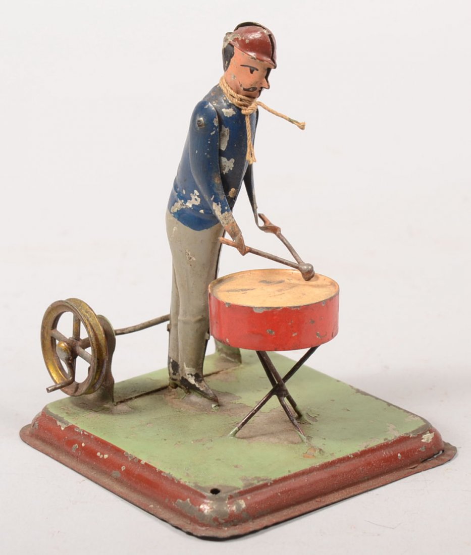 Painted Tin Man Beating on a Drum PTO Driven Toy.: Painted Tin Man Beating on a Drum PTO Driven Toy. Square tin base supports a pulley on a spindle with actuating arm to figure. Unmarked. 4 1/4"h.