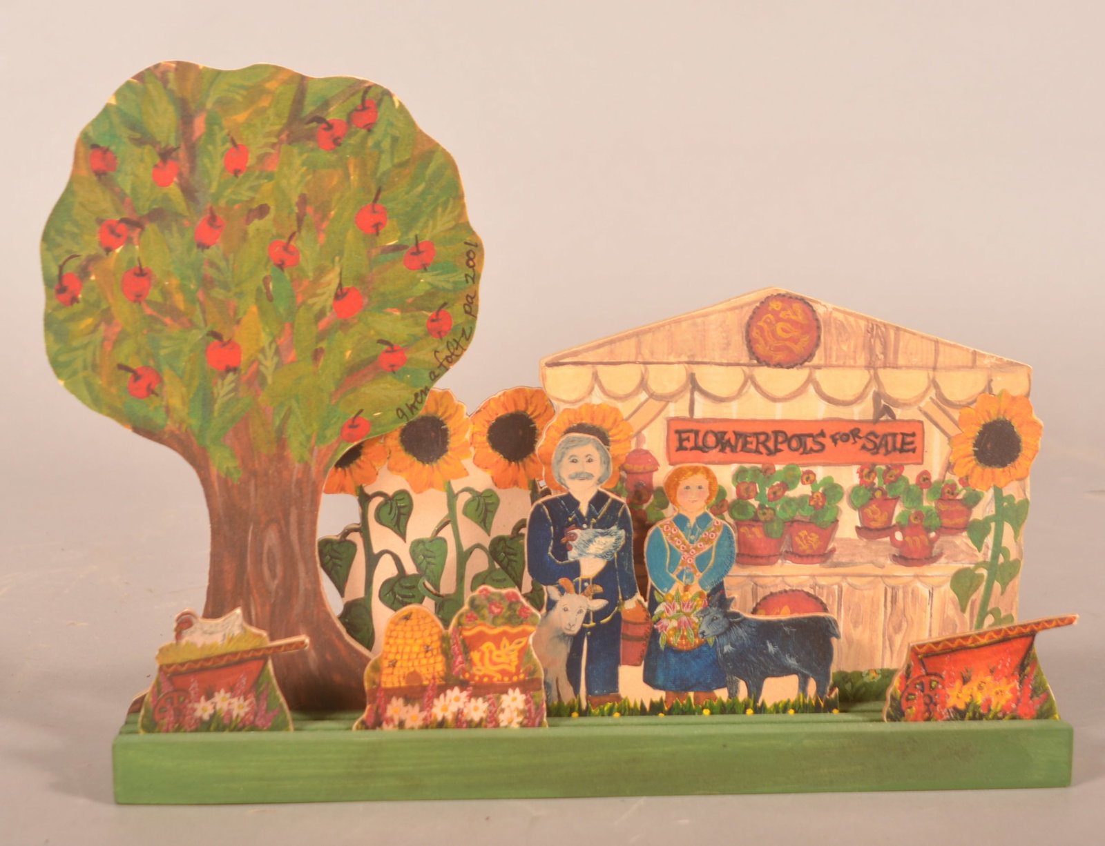 Gwen Foltz Folk Art Cut-Out Decorations: Original wood base with 7 wood and paper cut-out decorations, one signed "Gwen A Foltz Pa 2001". Decorations feature a bee skep, wheelbarrow, farmer's stand, a couple with animals, and sunflowers. 8 3