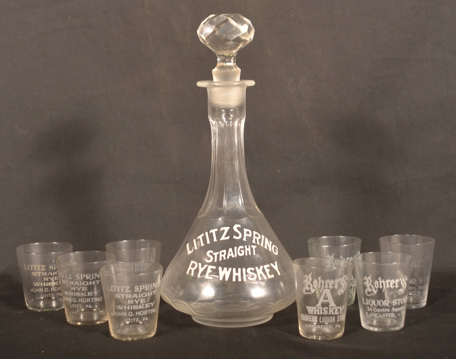 Lititz, PA Whiskey Bottle & Various Shot Glasses: Rare Lititz Springs Straight Rye Whiskey bar back bottle with white enamel painted letters and ground stopper. With 4 Lititz Springs John O. Horting shot glasses and 4 Rohrer's Whiskey Store Lancaster