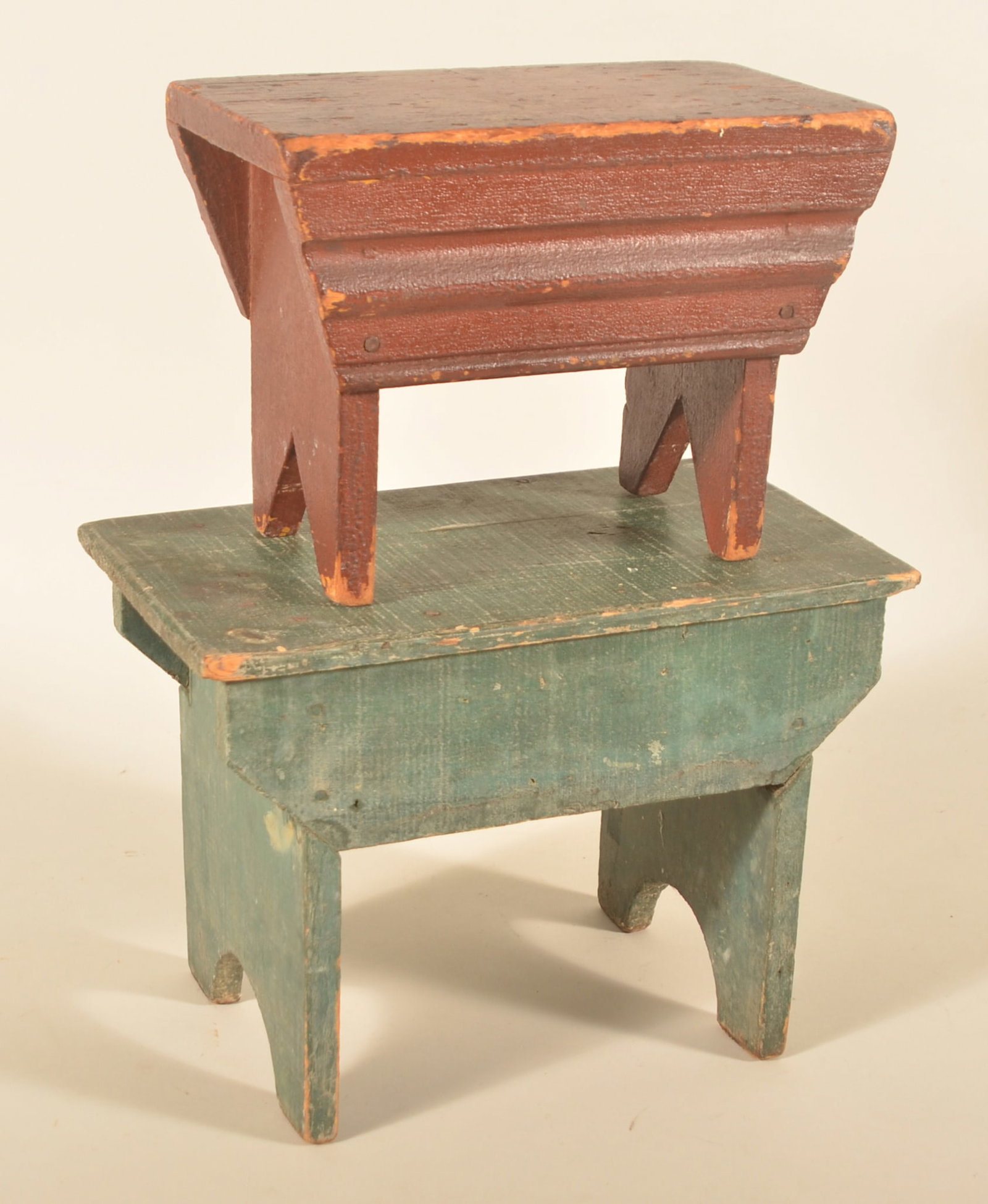 Two Painted Antique Pennsylvania Stools: Graduated stack of two painted stools. The smaller in an alligatored dark red paint with bootjack feet and shaped skirt on each side. Taller stool in a blue/green paint with half-moon cut-out feet. Ea