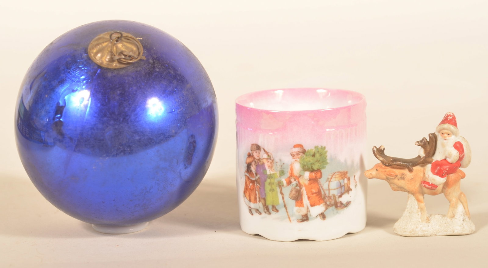 Antique Porcelain Santa Cup, Figural Santa & Kugel: Medium size, cobalt blue German Kugel with pressed tin cap. Small early-20th-century German child's porcelain cup with Santa bringing a tree to children. Small porcelain figural Santa riding a reindee