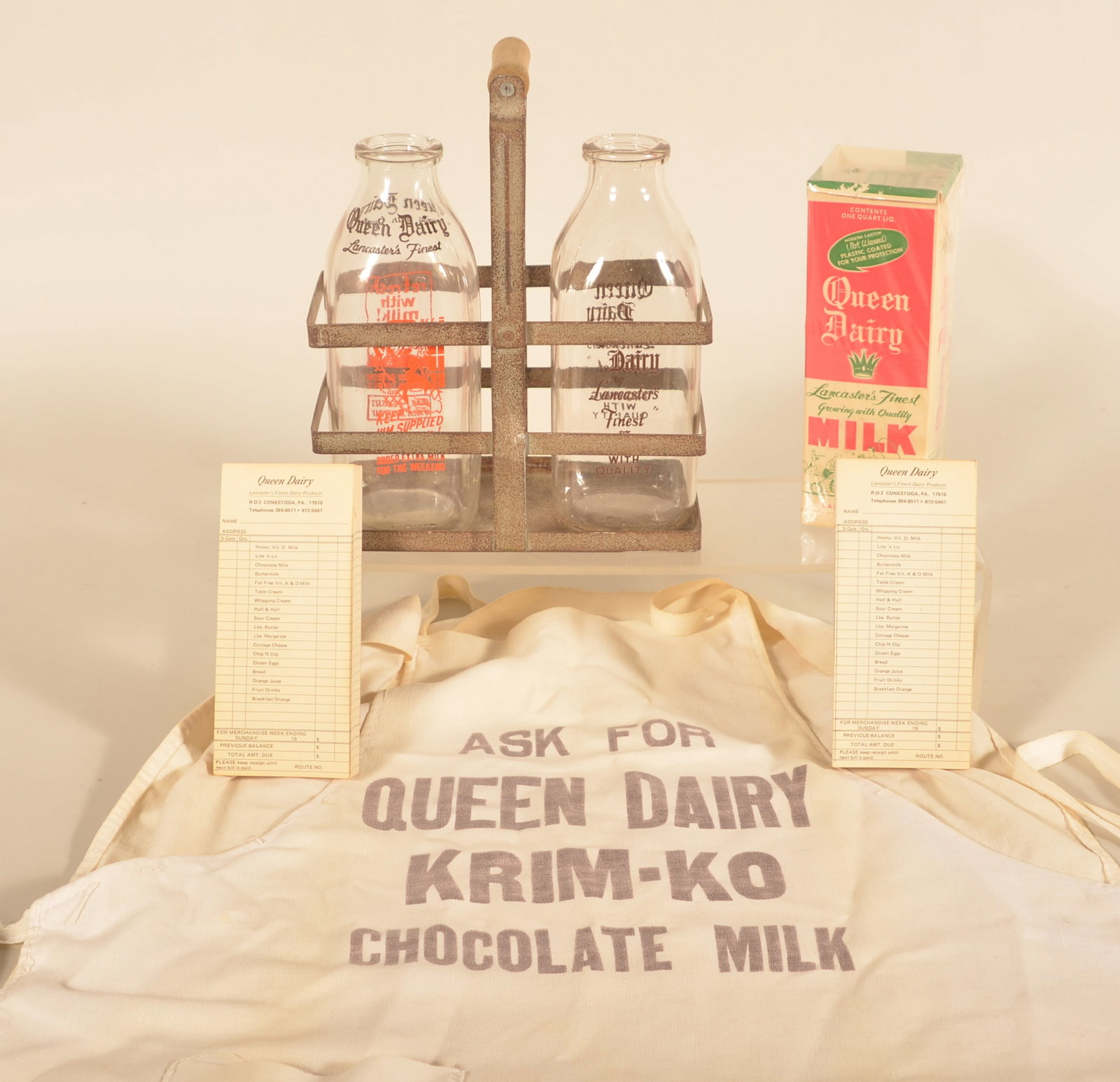 Queen Dairy Milk Bottles & Ephemera: Two Queen Dairy bottles in a vintage galvanized carrier, two original order forms, cardboard 1-quart milk container, and a printed server's apron. Nice group from Conestoga, Pa. Provenance: The collec