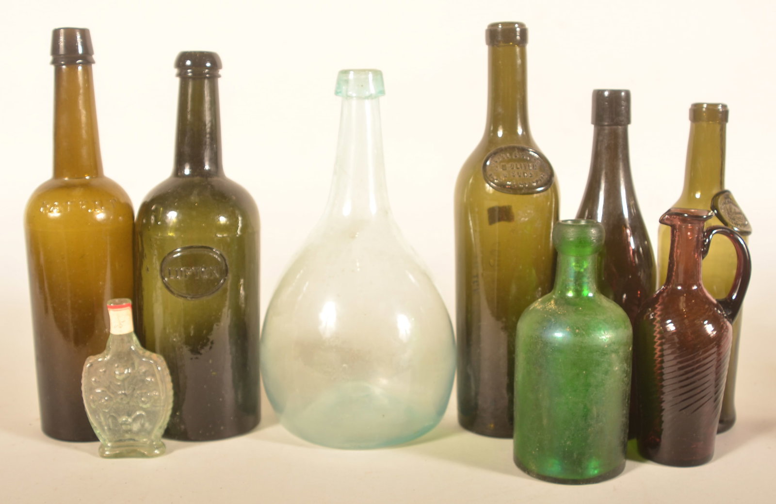 Collection of Antique Olive Oil & Spirits Bottles: Nine items in this collection including four dark green bottles, emerald green blob top bottle, amethyst bottle, embossed perfume bottle, amethyst blown glass pitcher and an open pontiled clear glass 