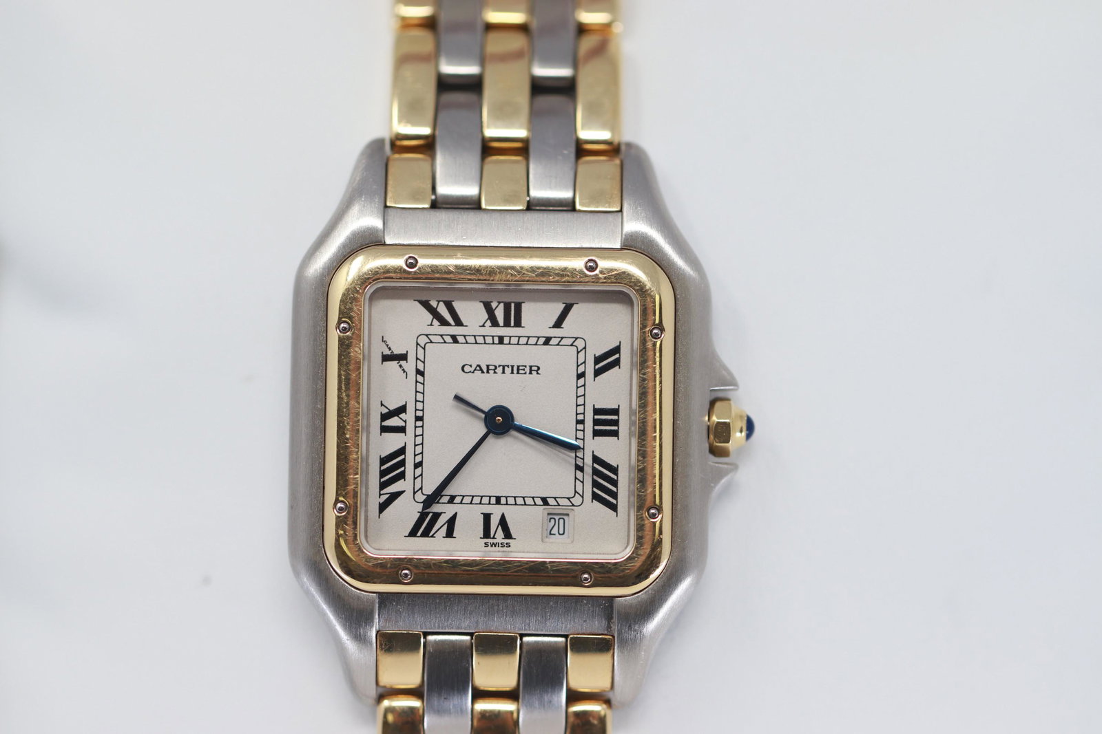 Cartier Panthere Steel & Gold Midsize 27mm Watch Ref.: A square 27 mm steel and gold case, featuring a white dial with Roman numerals, date aperture at 5 o'clock, surrounded by an 18k yellow gold bezel with a sapphire crystal and with a blue gem set, cabo