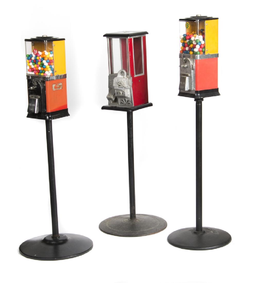 Three vintage gum ball machines on stands: A 'The Master' red and black enameled and glass 1 cent machine with turn mechanism and two red and yellow enameled and plastic 5 cent machines with turn mechanisms. Greatest height 46in.