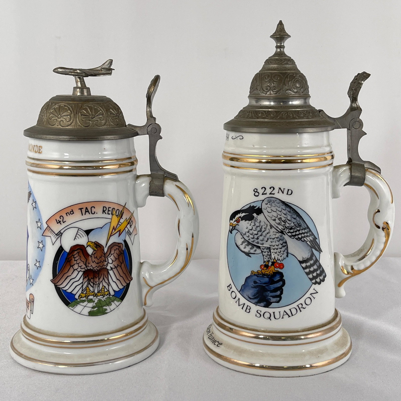 Two Lithophane Beer Steins with Pewter Lids: Circa 1950s. One Schmidt Koblenz "Spangdahlem Air Base, Germany" stein with Jetplane finial; and one 822nd Bomb Squadron stein; both with lithophane bottoms depicting seated nude females. Tallest 9.5"