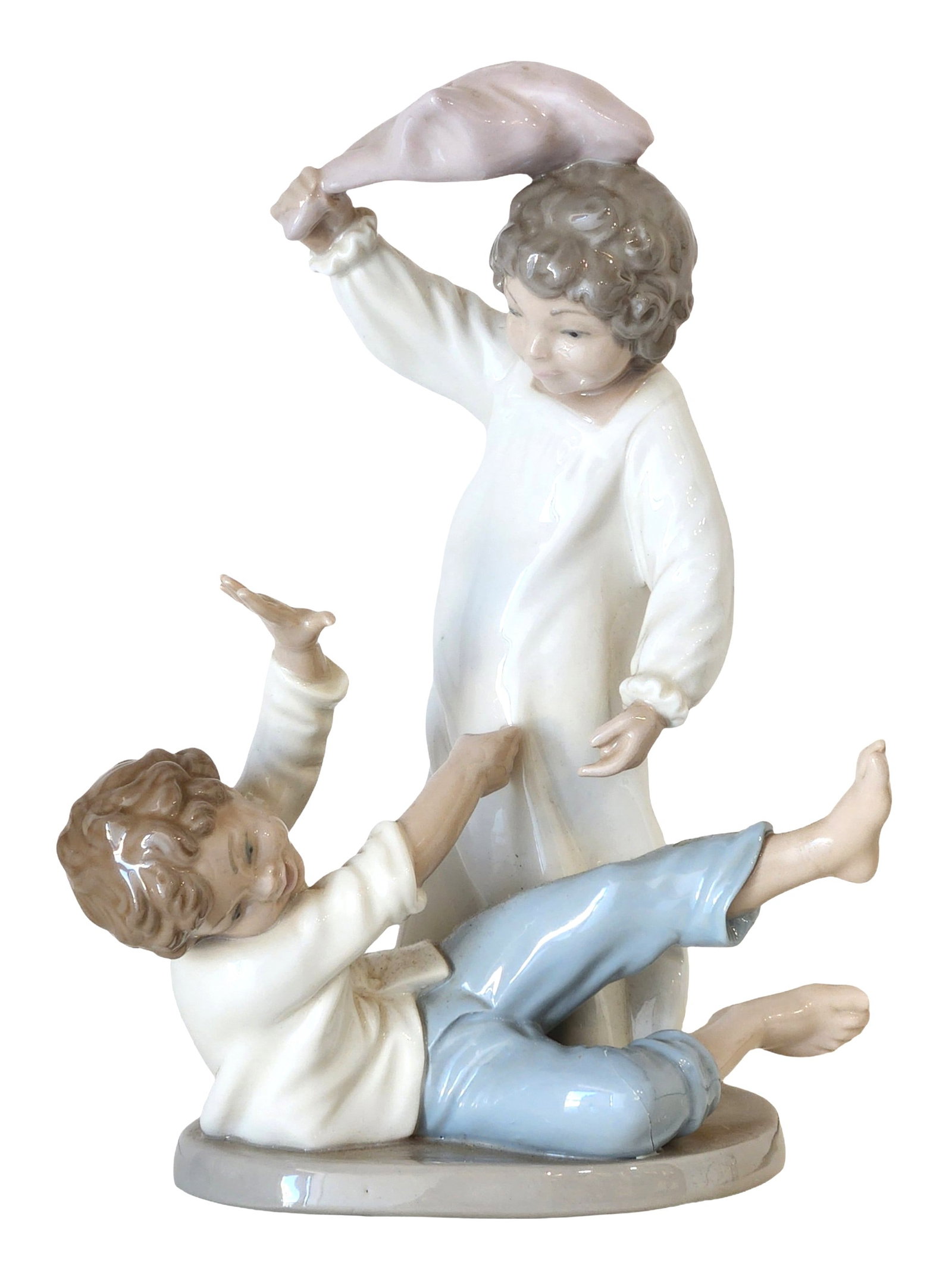 Lladro Porcelain Figurine " Two Children Playing with Pillow " Spain, 1997: Charming glazed porcelain figurine by Lladro depicting two young children engaged in playful interaction. One child stands in a long white nightgown, holding a soft pillow aloft, while the other recli