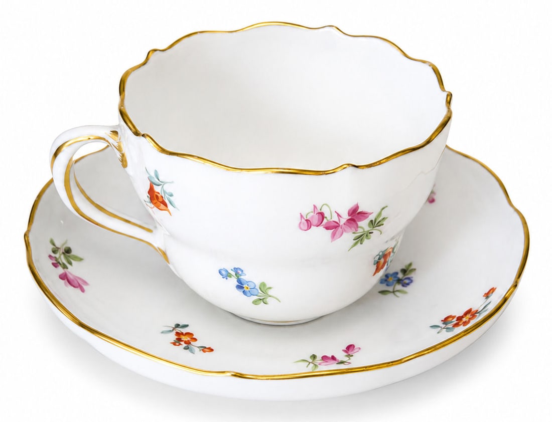 Meissen Porcelain Cup and Saucer with Hand Painted Floral Decoration: An elegant Meissen porcelain cup and matching saucer, finely decorated with delicate hand painted scattered floral sprigs in polychrome enamels. Both pieces feature softly scalloped rims accented with