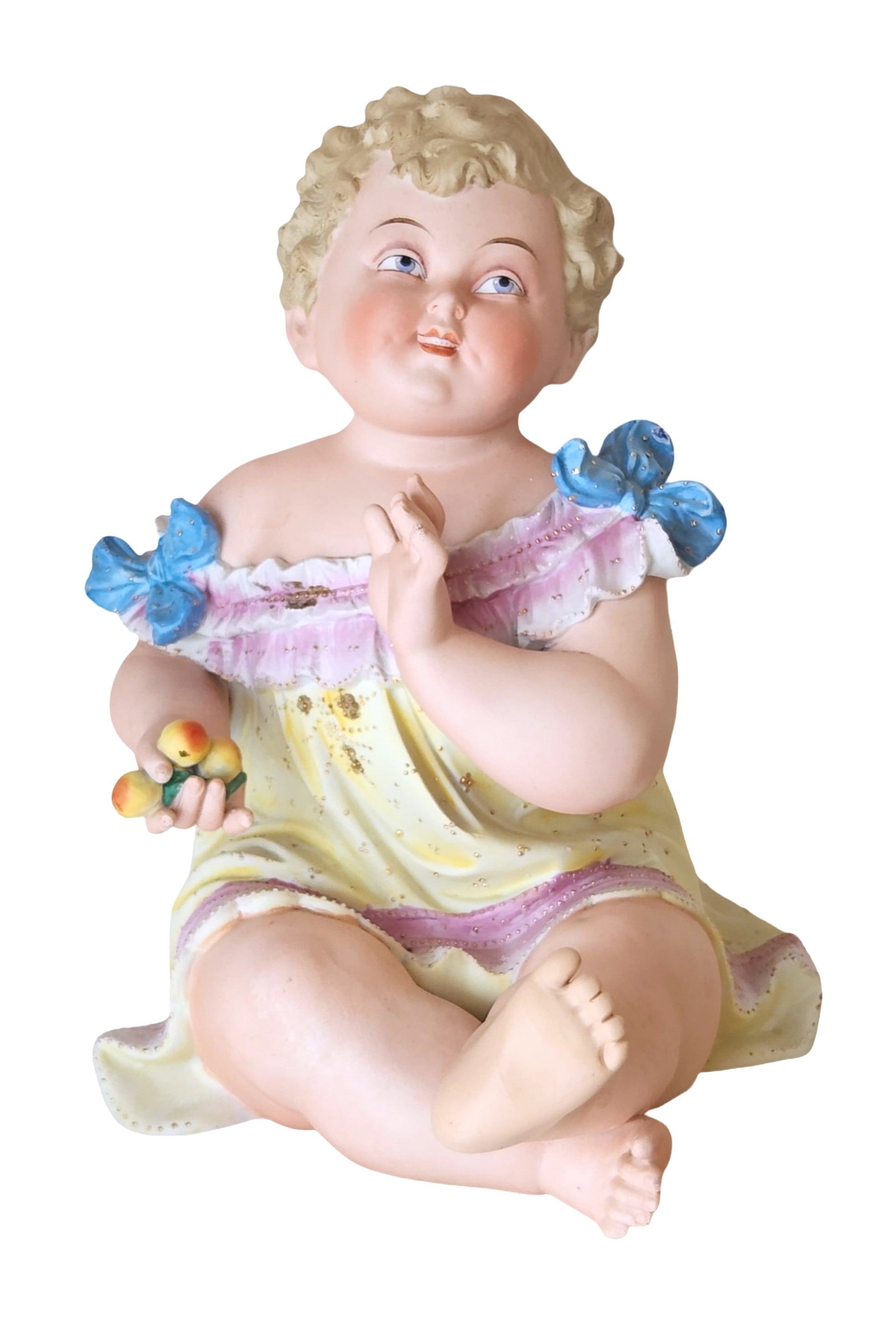 Antique Bisque Porcelain Baby with Fruit Piano Baby Figurine: Delightful antique bisque porcelain piano baby figurine, depicting a chubby child seated with one leg tucked under, wearing a ruffled pastel dress adorned with blue bows and holding a cluster of fruit