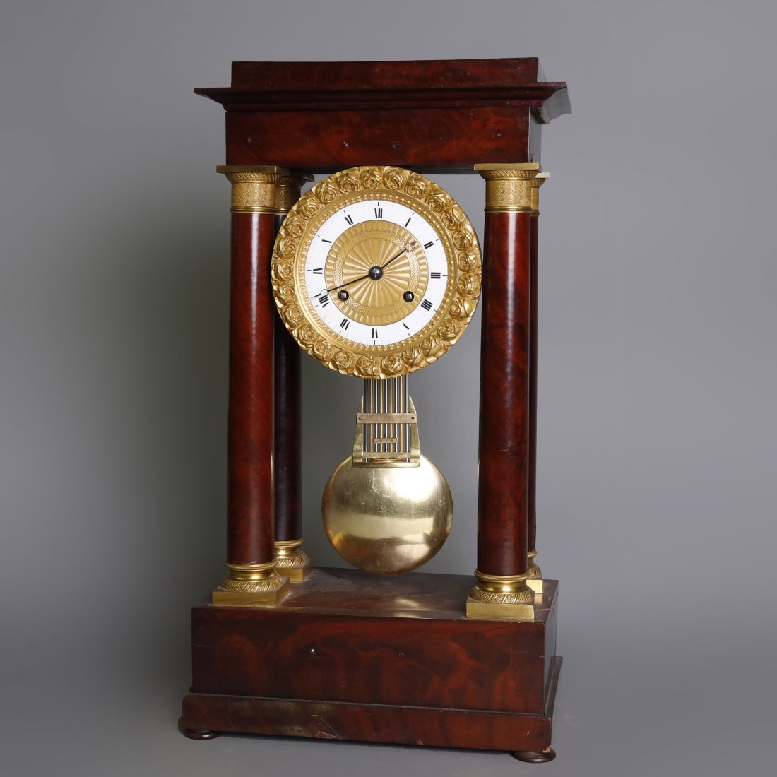 French Empire Mahogany & Ormolu Portico Clock, c1890: An antique French Empire portico clock offers flame mahogany case with full Corinthian column supports having bronze capitals and bases, face with cast floral Ormolu bordering surrounding Roman numera