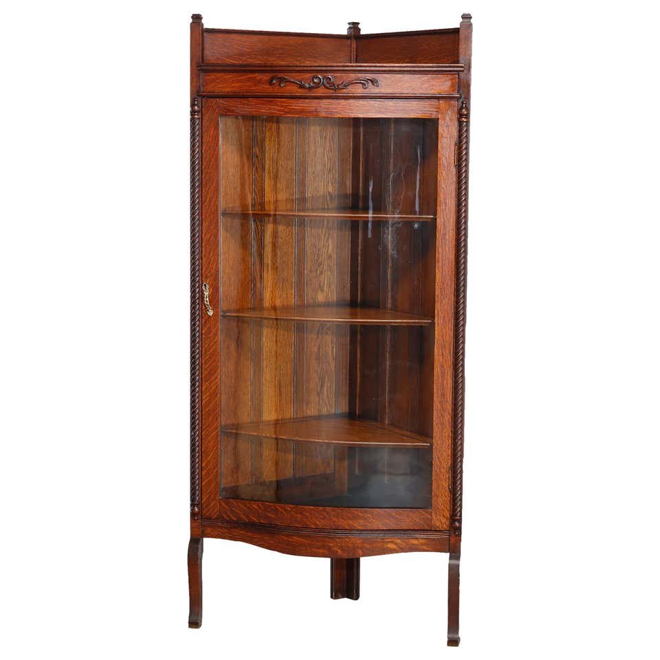 Antique RJ Horner Style Oak Corner China Cabinet, c1910: An antique corner china cabinet in the manner of R.J. Horner offers oak construction with upper backsplash surmounting carved foliate decoration and shelved case with bowed glass door flanked by rope 