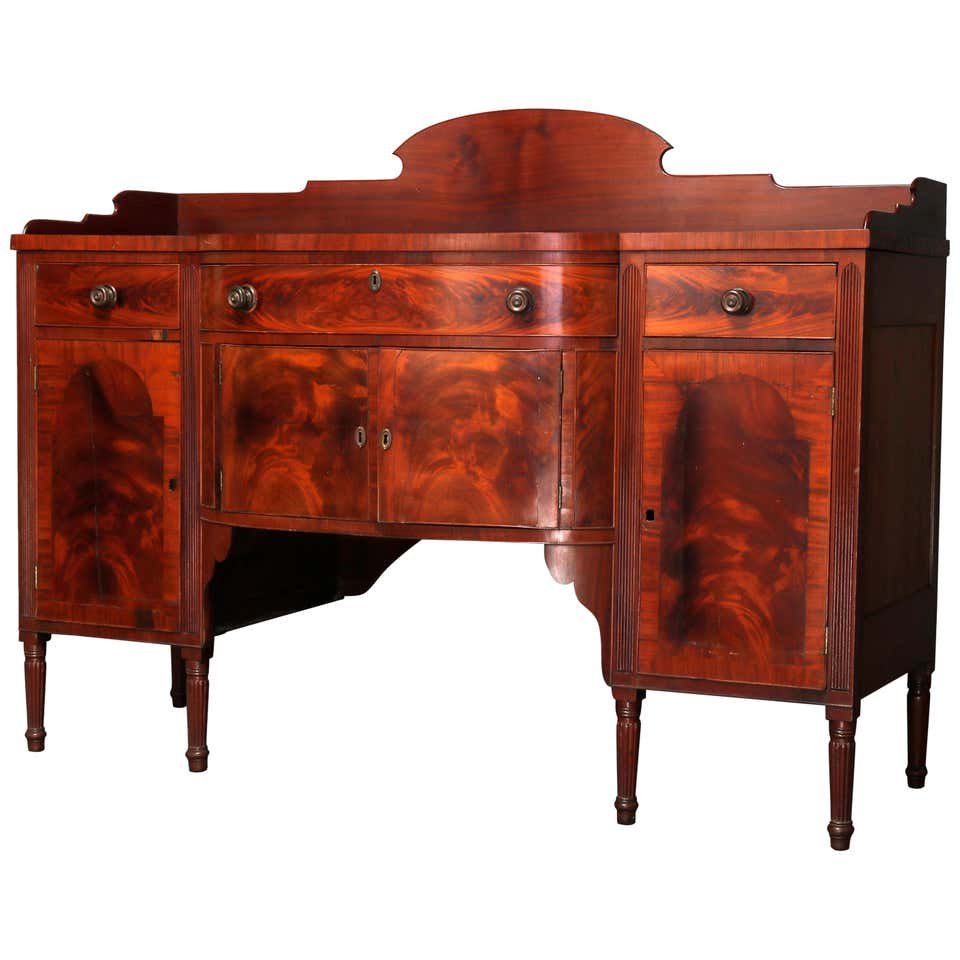 Antique Period Sheraton Flame Mahogany Sideboard,19th C: An antique period Sheraton sideboard offers flame mahogany construction with shaped backsplash surmounting case with bow front having frieze drawer flanked by two smaller and over double cross banded 