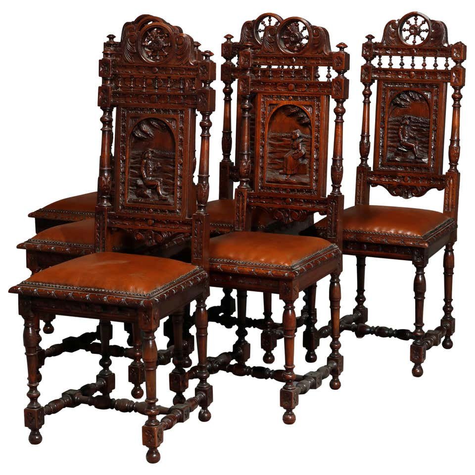 6 Continental Carved Genre Scenes Oak Dining Chairs: An antique set of six Continental dining chairs offers oak construction with wagon wheel form crest surmounting spindled and deeply carved backs with arch reserve having individual and different genre