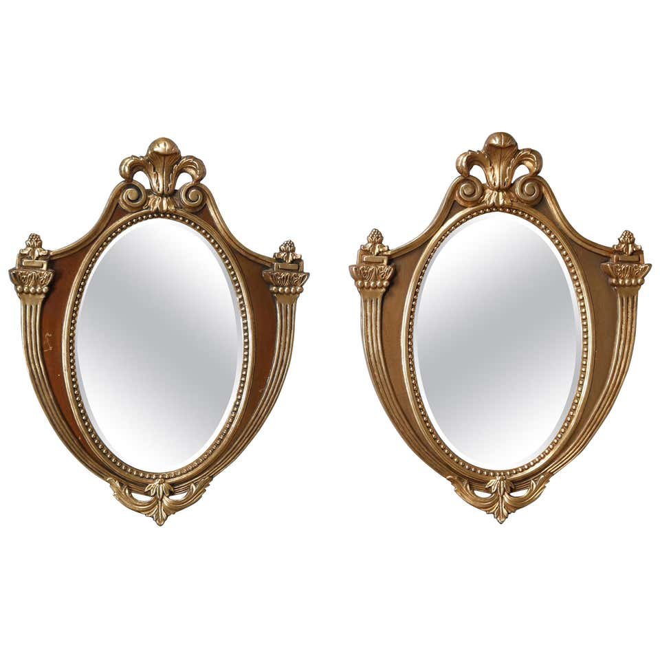 Pair French Parcel-Gilt Mahogany Shield Wall Mirrors: A vintage pair of French wall mirrors offer parcel-gilt mahogany frames in shield form with Fleur-de-Lis crest, beaded bordering, foliate elements and flanking stylized torchieres, circa 1940. Measure