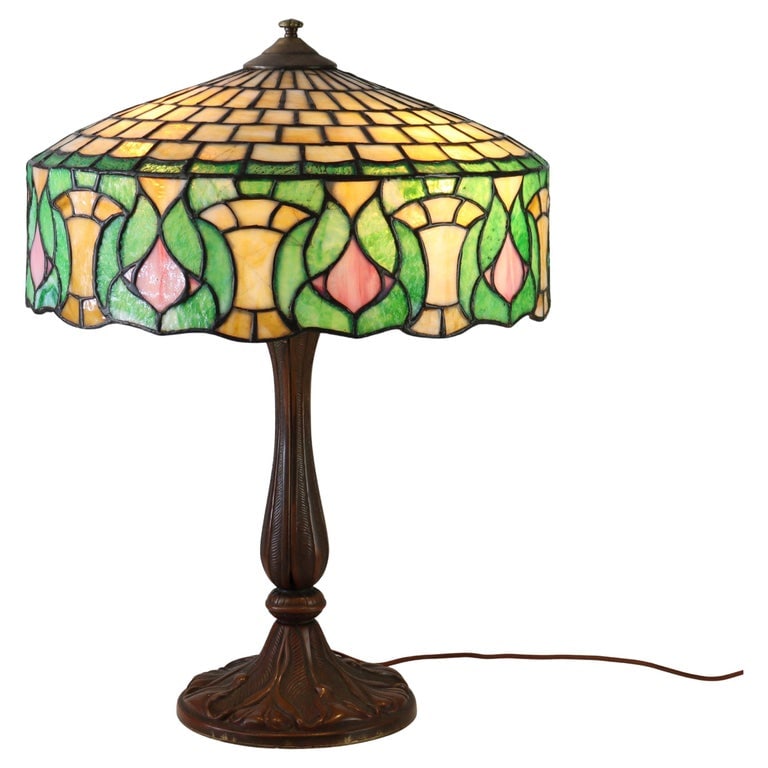 Whaley Art Nouveau Stained Leaded Glass Table Lamp with Bronze Base c1920: This Art Nouveau table lamp features a stained leaded glass shade with intricate floral and geometric patterns in vibrant hues of green, pink, and yellow. The lamp is supported by a bronze base, showc