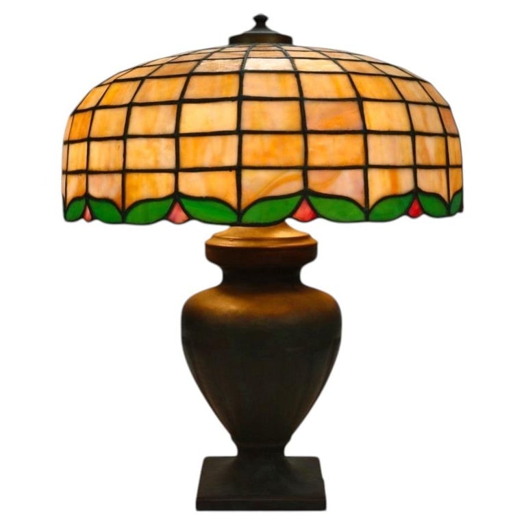Oversized Antique Handel Leaded Stained Glass Table Lamp With Bronze Base c1920: This oversized antique Handel table lamp features a leaded stained glass shade with a geometric grid pattern in warm amber and cream tones, accented by a border of green leaves and pink floral motifs.