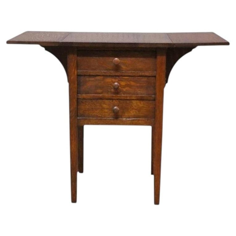 Stickley Form Arts & Crafts Mission Oak 3-Drawer Drop Leaf Stand C1910: This antique sewing stand, crafted in the Arts & Crafts Mission style, is constructed from solid oak with a rich, dark finish. Dating to approximately 1910, the stand features a rectangular top flanke