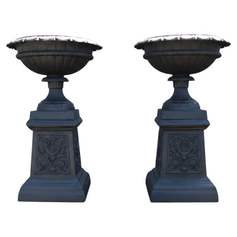 Pair Cast Iron Garden Urns With Stylized Floral Bases C1900: This offering presents a distinguished pair of antique cast iron garden urns, dating to circa 1900. Crafted from robust cast iron, these planters exemplify classical design influences, making them ide