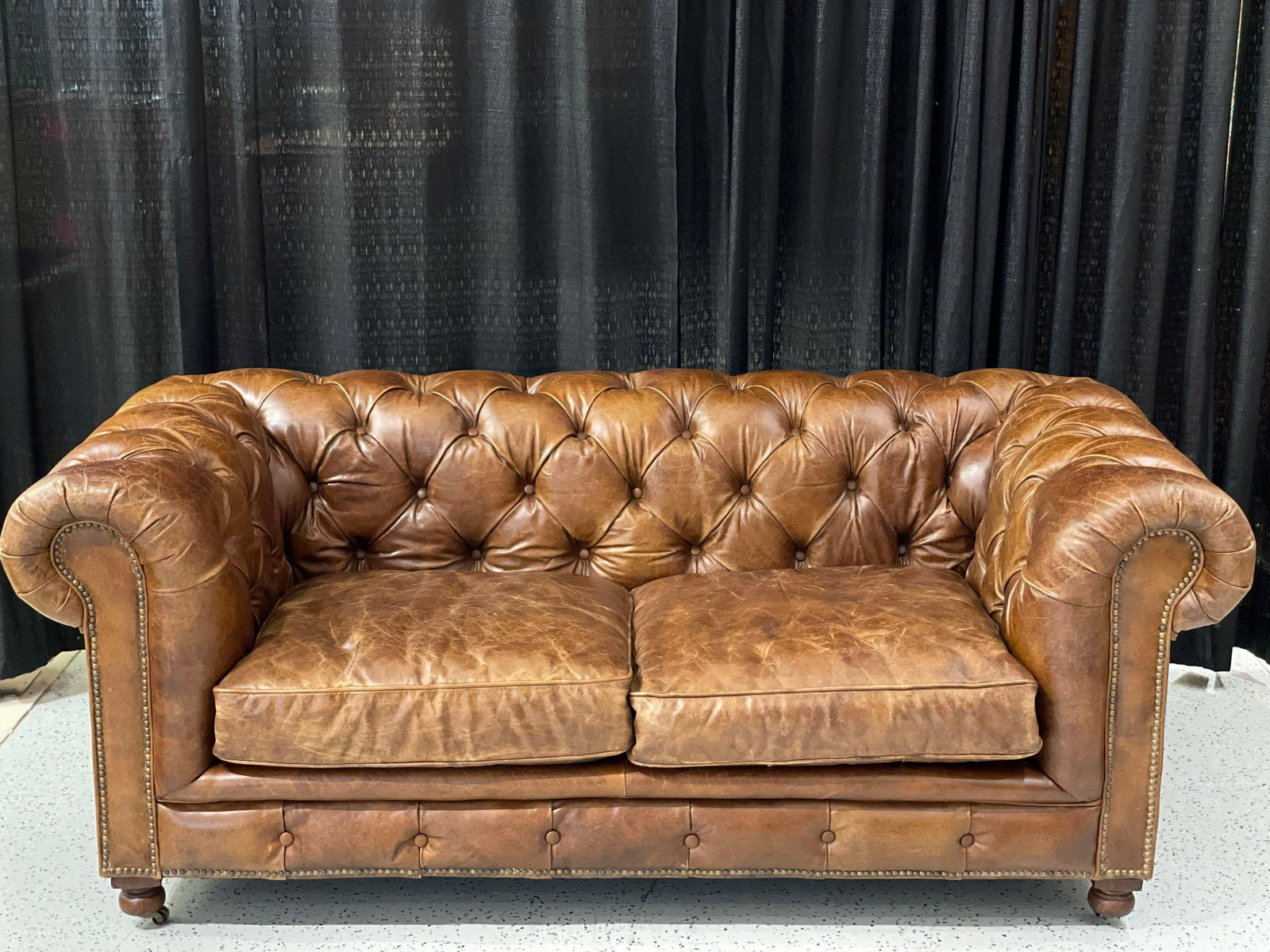 Halo Limited Tufted Leather Love Seat: Dark brown tufted leather love seat by Halo Asia Limited. Features a tufted low back design, studded accents, and is equipped with castors. One of the castors has broken off. The leather does show som
