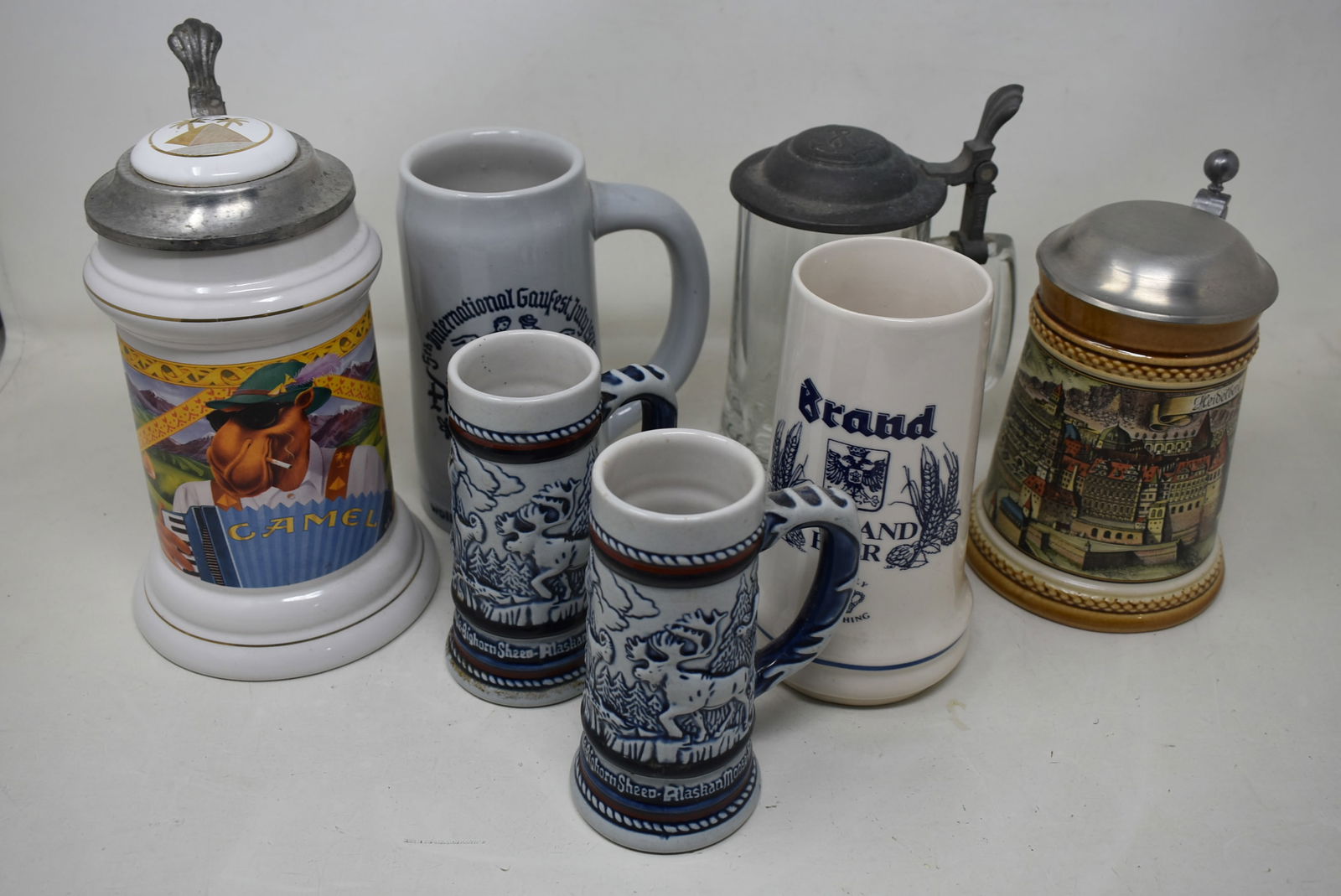 Group of 7 Beer Steins: A Lot of 7 Beer Steins including: 2 Avon, 1 Ceramarte Alpenland, 1 Brand Holland, 1 Camel, 1 Beyer Heidelberg Lidded and 1 Glass Stein with Ship Scene "Columbia 1901". Good condition.