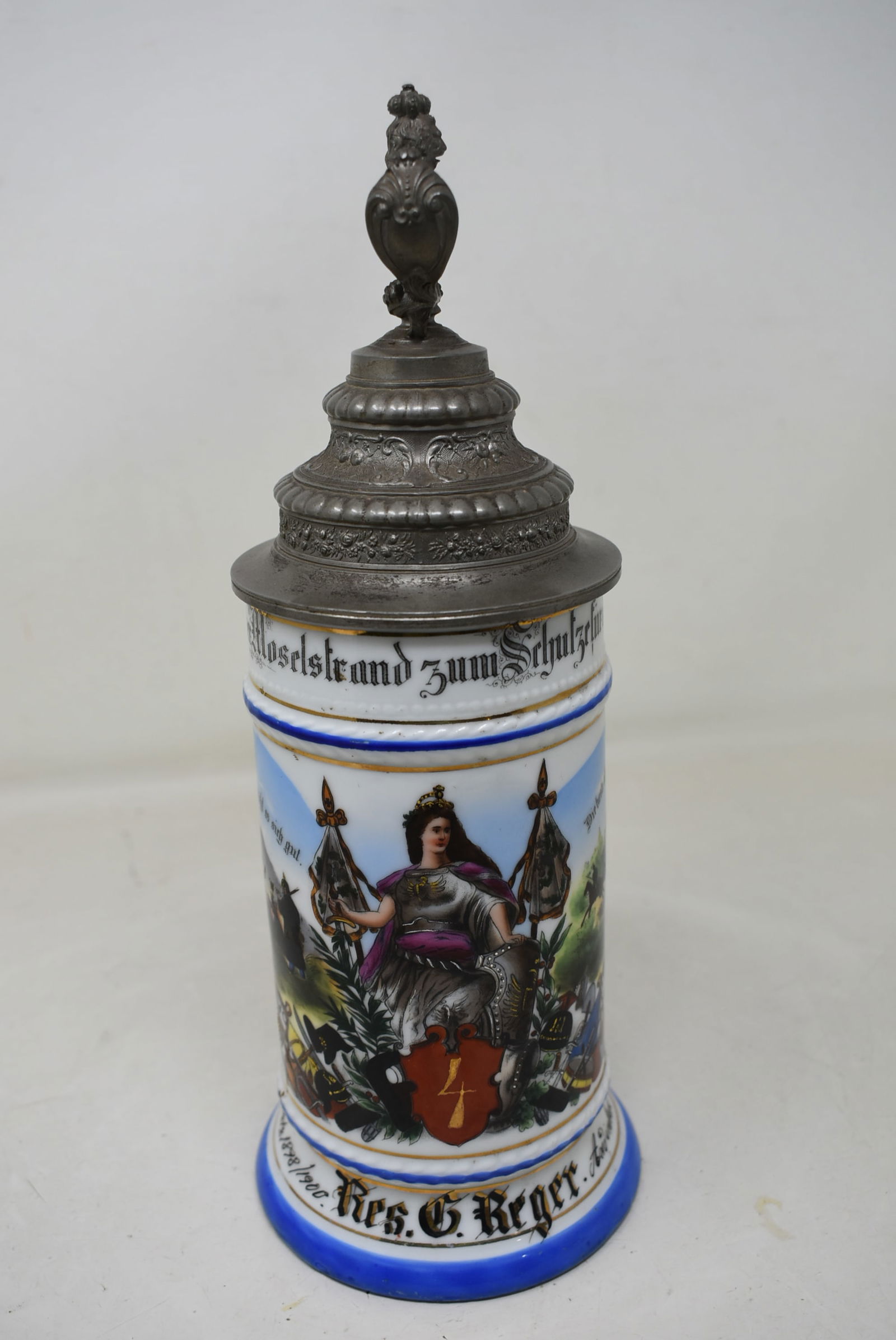 1/2 Liter German Regimental Porcelain Beer Stein 1898-1900: German Regimental Beer Stein 1898/1900, "Res. G. Reger", Battle Scenes and Female Figure. Lion Finial and Crown Thumb lift. Interior rim has a small chip, repair inside of lid at strap, strap has play
