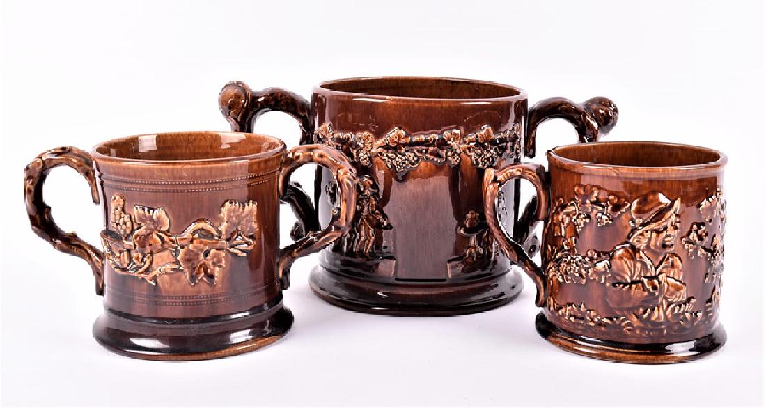 A collection of three Staffordshire treacle-glazed frog: A collection of three Staffordshire treacle-glazed frog mugs comprising two loving cups and one with single handle, each with applied sprigs, the largest 16 cm high x 32 cm wide. (3)