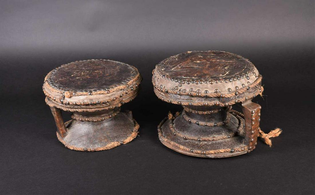 A near pair of West African hardwood drums, of shallow: A near pair of West African hardwood drums, of shallow dumb bell form, the skins secured by domed copper studs, each with a band of incised decoration, further decorated with bands of rope secured by