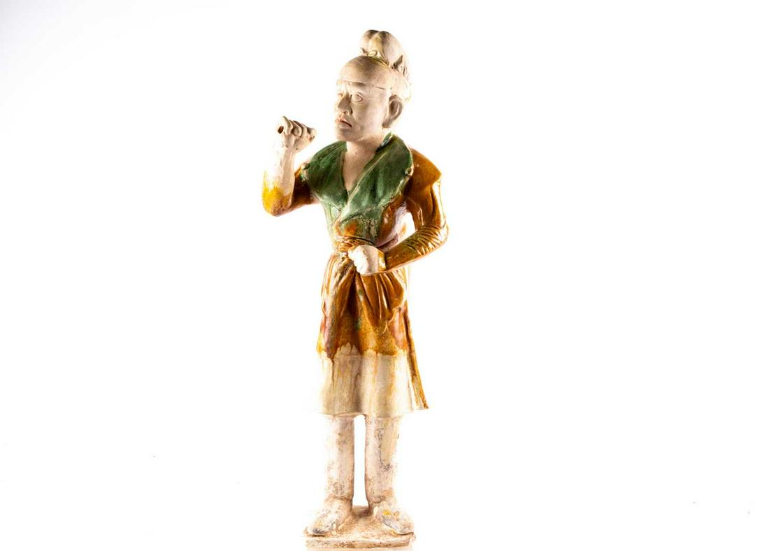 A Chinese Sancai glazed pottery figure of a Western: A Chinese Sancai glazed pottery figure of a Western groom, Tang dynasty, modelled standing, his face with a concentrated expression, a splash of yellow glaze to his cap, his left arm raised as if to h