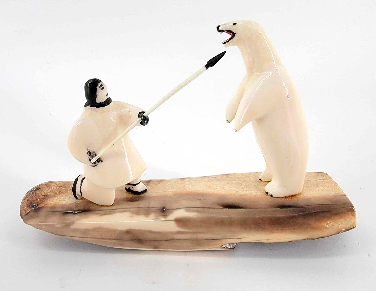 Walrus Tusk Polar Bear & Hunter Carving: A tusk sculpture with scrimshaw details depicting an Alaska Native man fighting off a standing polar bear with a spear, mounted on a fossilized tusk base and signed. Measures approximately 6.5" x