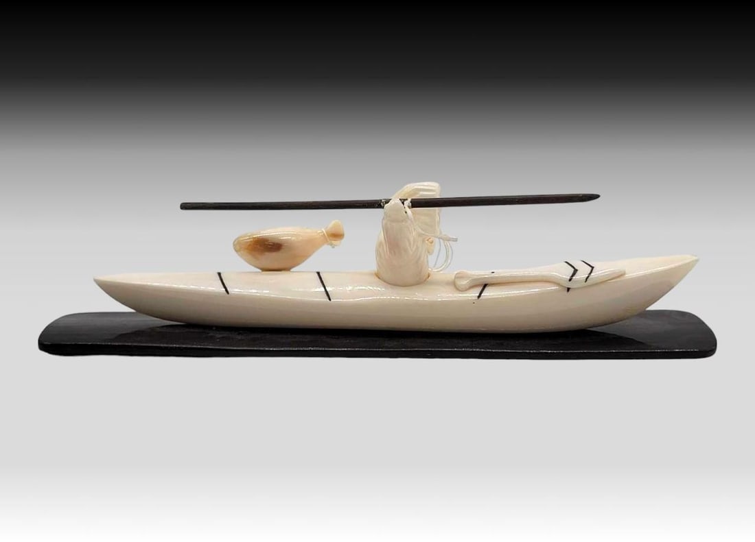 Walrus Tusk Seal Hunter w/ Kayak & Baleen Spear - Allen Kulukhon: A carved walrus tusk seal hunter sitting within his tusk kayak, a tusk paddle perched upon the bow and his tusk sealskin air bag upon the stern. A baleen spear is within his right hand and his left gr