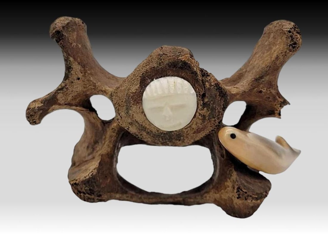 Fossilized Vetebra & Walrus Tusk Sculpture - Charles Slwooko: A fossilized vertebra with inset walrus tusk carved face, inserted within the spinal cord cavity; a fossilized tusk fish with inset baleen eye is mounted upon the side in a swimming pose. A portion of