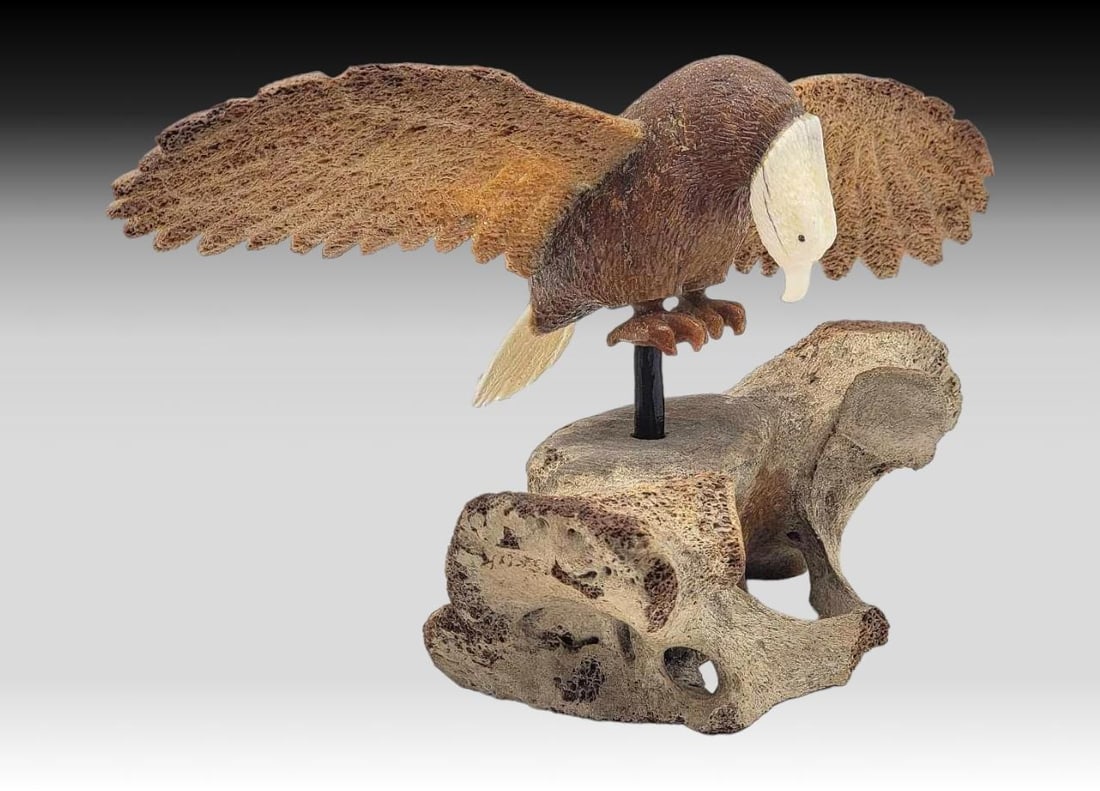 Walrus Tusk & Fossilized Whale Bone Bald Eagle on Walrus Vertebrae: A meticulously carved walrus tusk depicting the body of a bald eagle, accented with fossilized whale bone wings and inset baleen eyes. The majestic eagle rests upon a walrus vertebra via a baleen rod,