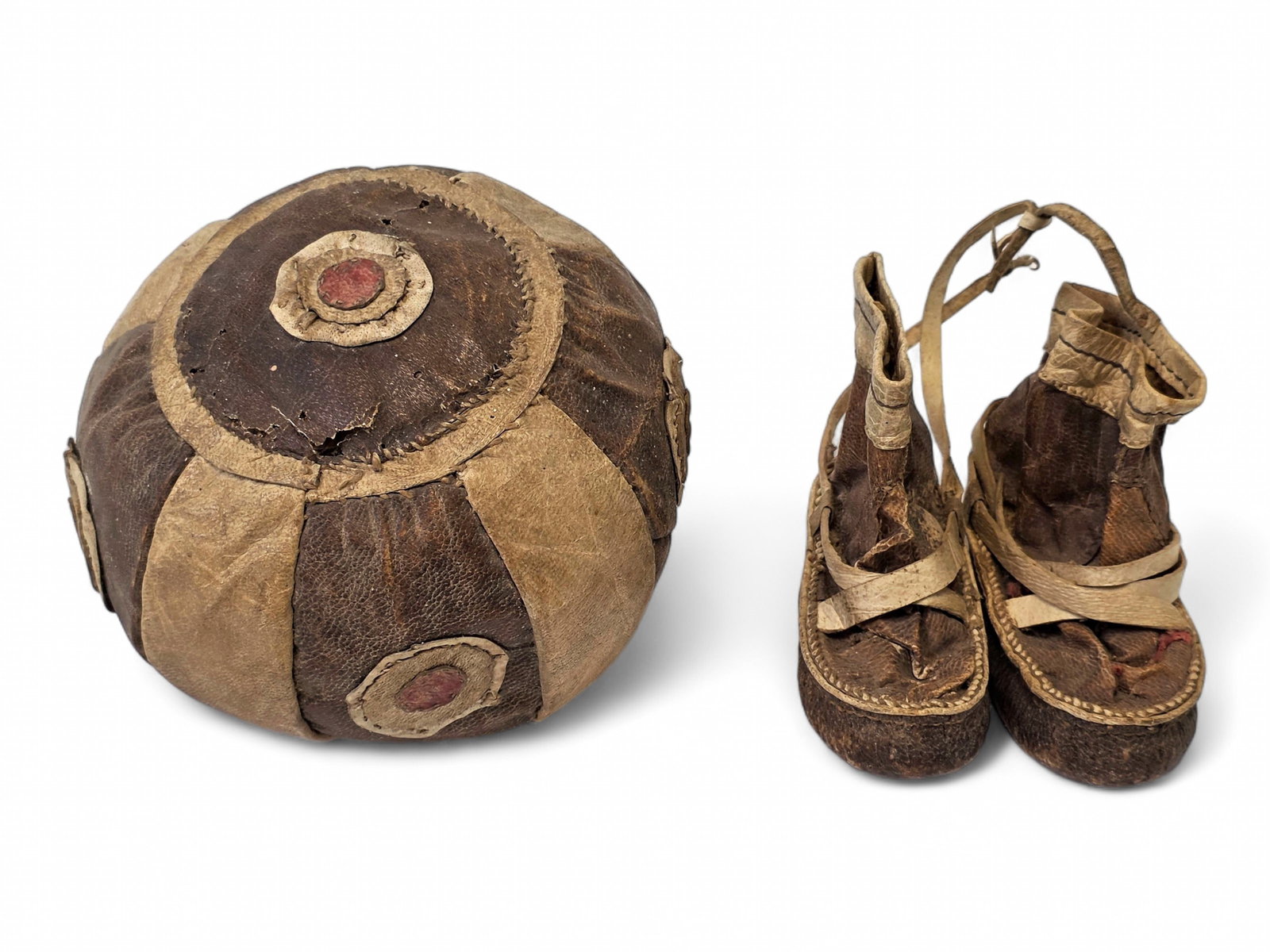 Antique Yup'ik Child-Sized Mukluks & Game Ball: A vintage Yup'ik game ball primarily crafted from two shades of brown hide- likely walrus/seal, and featuring concentric circle motifs terminating in a red dot. Also includes a pair of vintage child-s