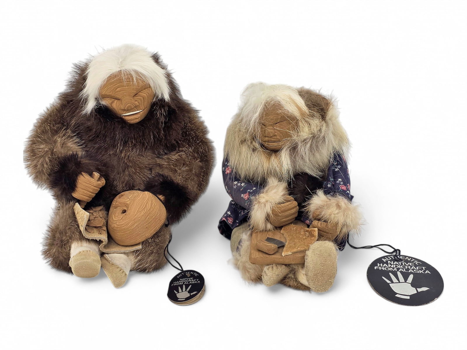 Yup'ik Story Dolls- Margaret "Penni" A. Konukpeok: Two Yup'ik story dolls, each hand-crafted with carved cottonwood faces and hands, hide mukluks, and fur hair, and both in seated positions. One doll is attired in a fur parka and is in the process of
