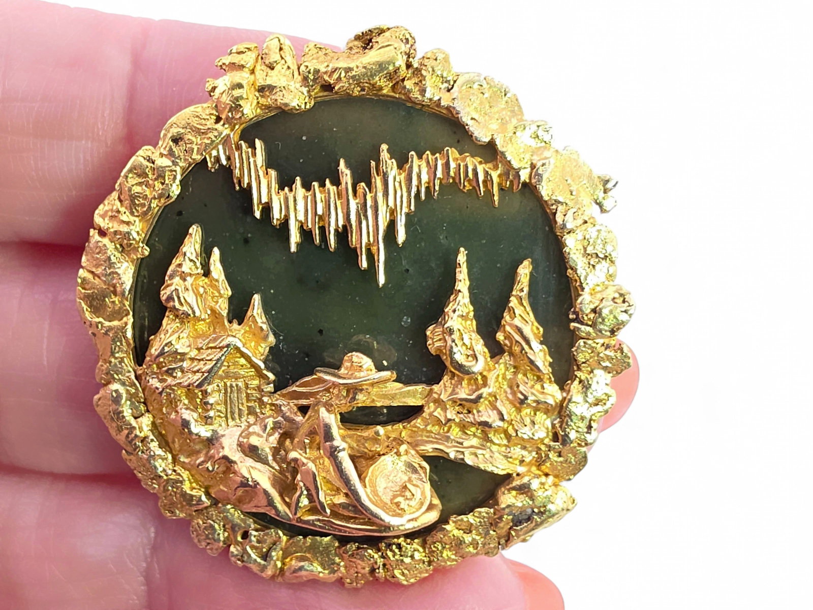 10K Gold, Alaskan Gold Nugget & Jade Prospector Aurora Brooch: A finely executed 10-karat yellow gold and nephrite jade brooch celebrating Alaska's mining heritage. The circular plaque is centered by a polished dark green nephrite jade disk framed by a rugged bor