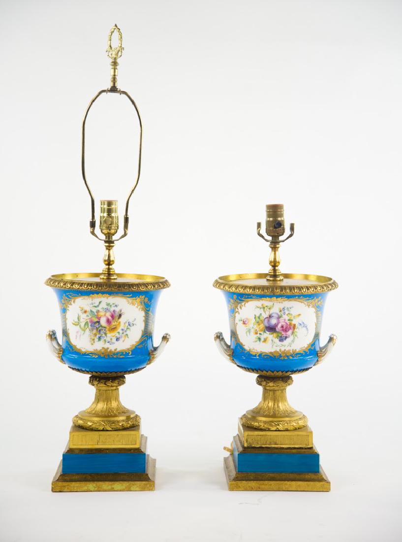 Pair of 19th Century Sèvres Celeste Blue Urn Lamps: A pair of mid-19th century porcelain French Dore Bronze Ormolu Urn Lamps having a square pedestal base. Egg and Arrow trim around top edge and hand painted playful bucolic period scene and floral moti