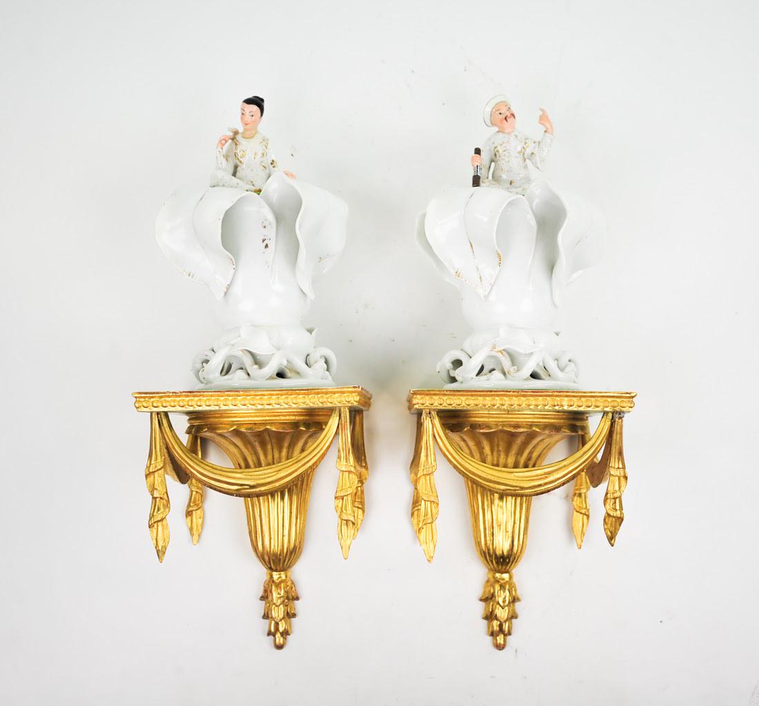 Pair of Antique Sconces: A pair of antique sconces. Each sconce features a giltwood base decorated with carved drapes. Atop each base is a porcelain figure, one male and one female, emerging from a white lily. They are unmark