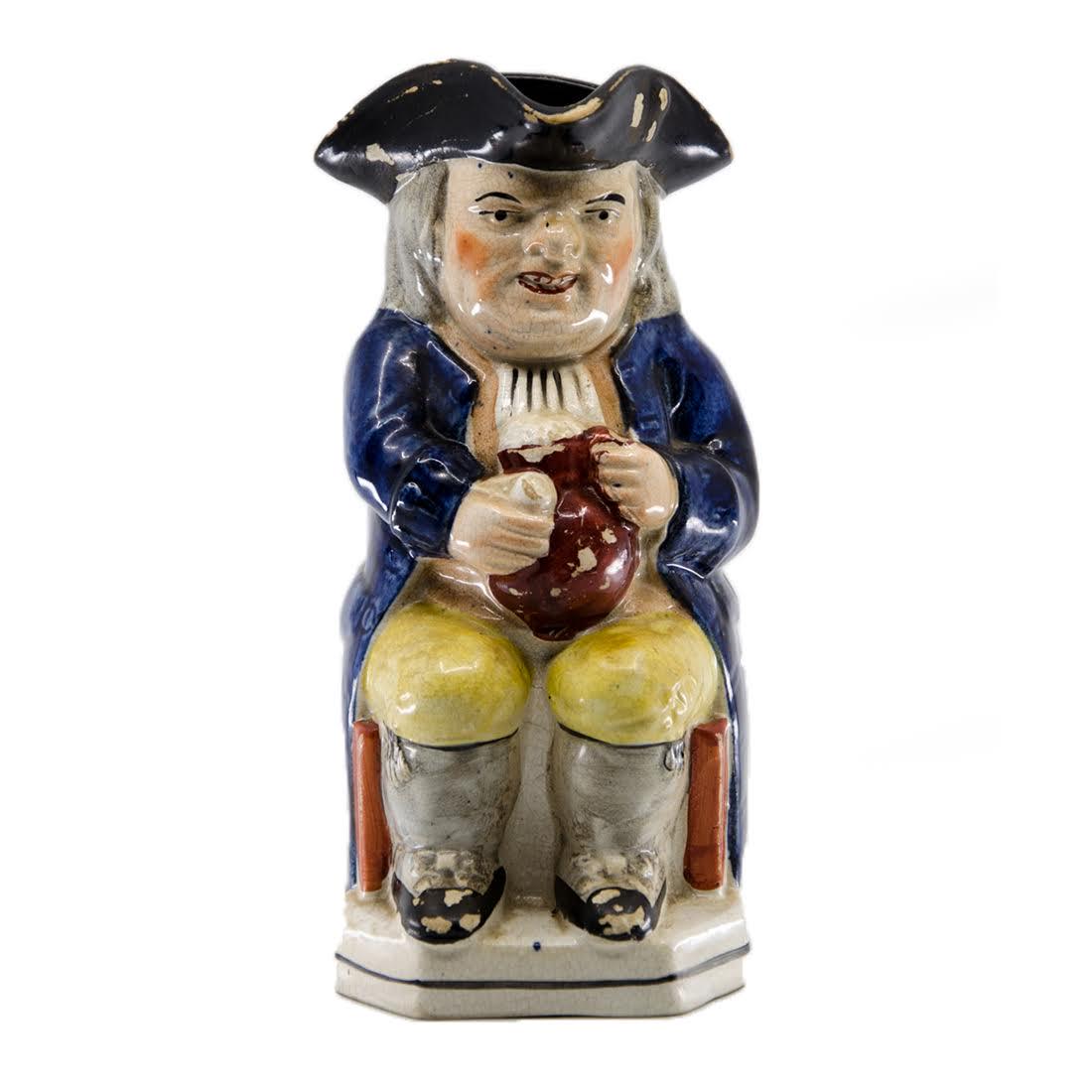 Antique Majolica Quaker Man Toby Jug: An antique majolica toby jug. This piece takes the form of a seated quaker man with yellow pants, grey socks, black shoes, an orange and white shirt, blue jacket, and black hat. He is holding a red ju