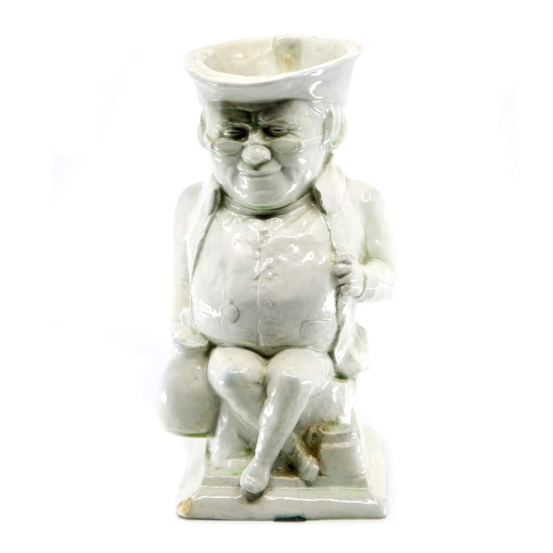 Antique White Glaze Old Man Toby Jug: An antique white glaze toby jug. This piece takes the form of a seated old man. His legs are crossed and he is holding a jug in his right hand. This piece is marked with an "F" on the interior. Dimens