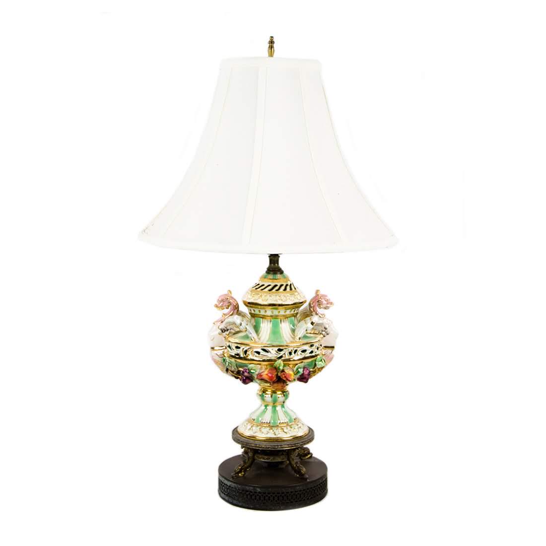 Antique Capodimonte Lamp with Silk Shade: An antique Capodimonte porcelain lamp with a silk shade. This piece features nude mastheads at each end, fruit appliques around the body, and gilt detailing throughout. It is unmarked. Dimensions: 29.