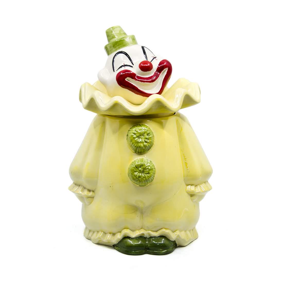 Metlox Poppy Trail Ceramic Clown Cookie Jar: A vintage Metlox Poppy Trail ceramic cookie jar. This piece takes the form of a laughing clown. He is wearing white and red face makeup, a yellow and green robe, and a green hat. It is marked "Made in